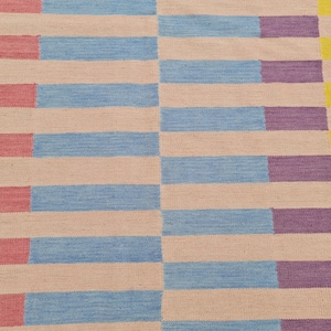 May include: A woven rug with a geometric pattern. The rug features alternating horizontal stripes of light pink, light blue, and purple, with yellow and pink vertical stripes on the edges. The rug is made of a textured material.