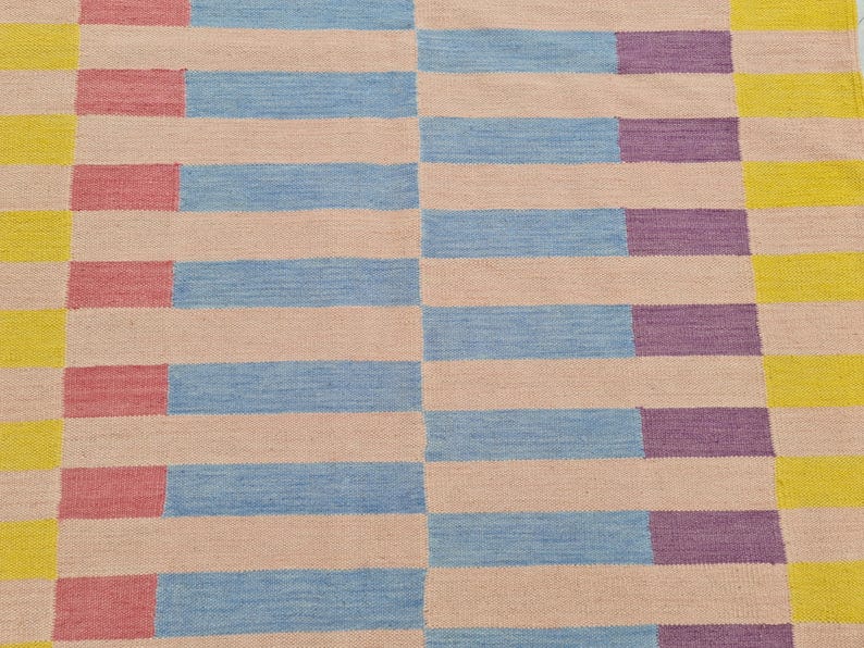 May include: A woven rug with a geometric pattern. The rug features alternating horizontal stripes of light pink, light blue, and purple, with yellow and pink vertical stripes on the edges. The rug is made of a textured material.