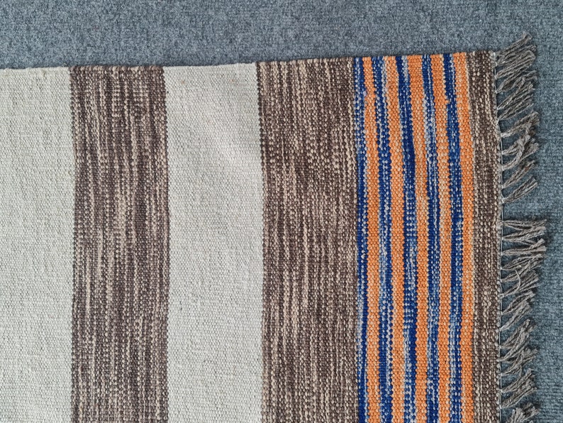 May include: A woven rug with vertical stripes in beige, brown, orange, and blue. The rug has a fringed edge and a textured appearance. The color scheme is neutral with a pop of color from the stripes.