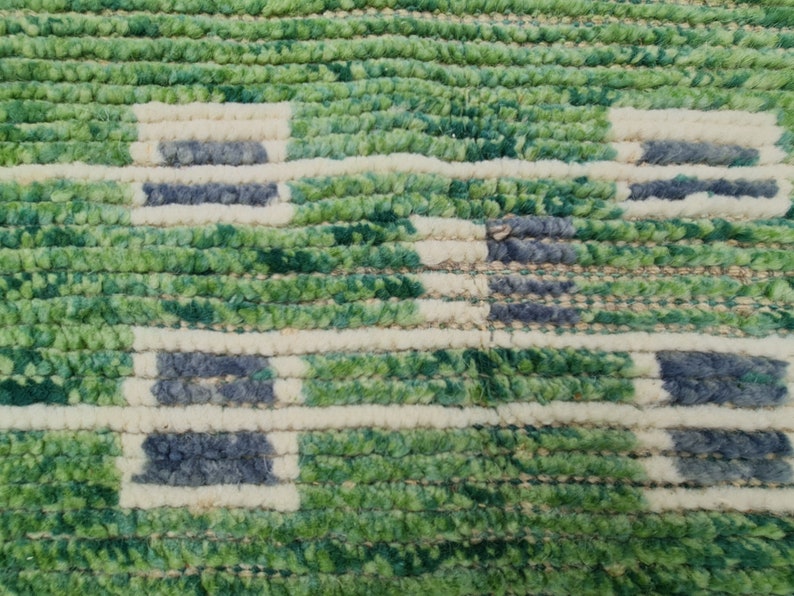 Puede incluir: Primer plano de una alfombra texturizada con un patr&oacute;n geom&eacute;trico repetitivo. La alfombra presenta una combinaci&oacute;n de colores verde, blanquecino y azul oscuro. El dise&ntilde;o incluye formas rectangulares y l&iacute;neas horizontales.