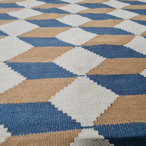 May include: A woven rug with a geometric pattern of diamonds in white, blue, and tan. The diamonds create a three-dimensional effect. The rug is made of a textured material, likely natural fibers.