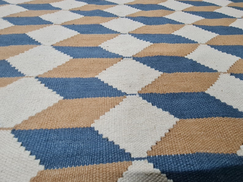 May include: A woven rug with a geometric pattern of diamonds in white, blue, and tan. The diamonds create a three-dimensional effect. The rug is made of a textured material, likely natural fibers.