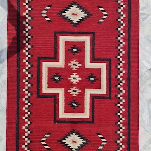 Pode incluir: Um tapete retangular vermelho com um padrão geométrico. O tapete apresenta um design central em forma de cruz em bege e preto, rodeado por motivos de diamante e seta. A borda tem um padrão repetitivo em preto e branco.