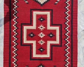 May include: A rectangular red rug with a geometric pattern. The rug features a central cross-like design in beige and black, surrounded by diamond and arrow motifs. The border has a repeating black and white pattern.