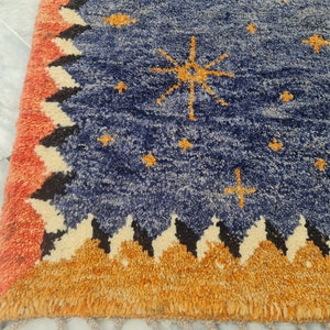 May include: A blue rug with a celestial design. The rug features a navy blue field with gold star and sunburst patterns. The border has a repeating pattern of orange, black, and cream zigzags, with a fringed edge in orange.