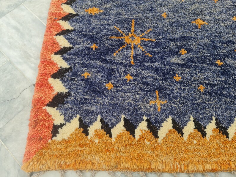 May include: A blue rug with a celestial design. The rug features a navy blue field with gold star and sunburst patterns. The border has a repeating pattern of orange, black, and cream zigzags, with a fringed edge in orange.