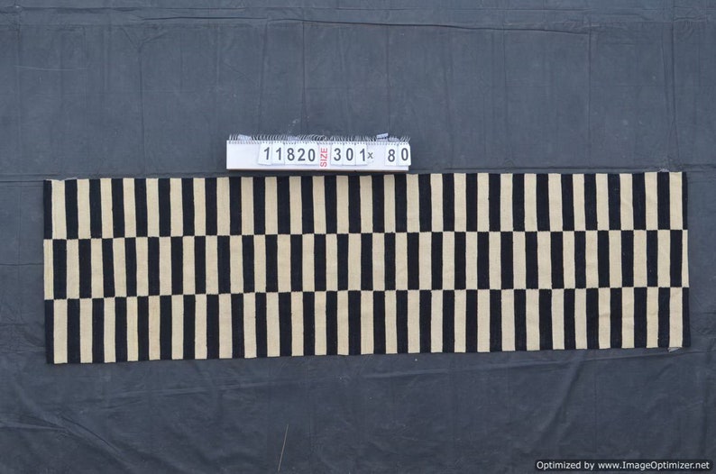 May include: A rectangular rug with a repeating pattern of vertical black stripes alternating with beige stripes. The rug is approximately 30 inches long and 80 inches wide. The rug is made of a woven material.