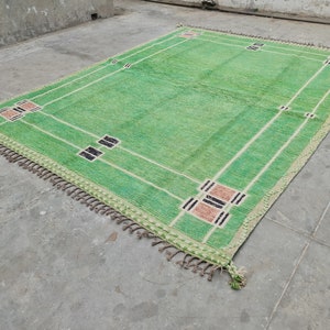 May include: A rectangular green rug with a geometric design. The rug features a light green field with white lines and decorative squares in black and tan. The edges are trimmed with a fringe.