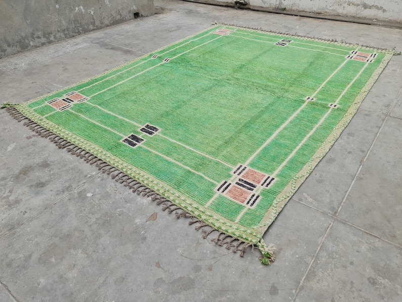 May include: A rectangular green rug with a geometric design. The rug features a light green field with white lines and decorative squares in black and tan. The edges are trimmed with a fringe.
