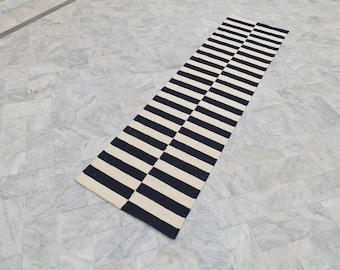 May include: A rectangular rug with a bold striped pattern in navy blue and cream. The rug is made of a woven material and is placed on a light gray tiled floor. The stripes run lengthwise across the rug.