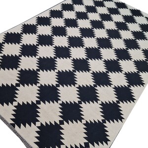 Puede incluir: Una alfombra rectangular con un patr&oacute;n repetitivo de cuadrados negros y crema. Los cuadrados tienen un borde dentado, casi en forma de estrella. La alfombra est&aacute; hecha de un material tejido y tiene una paleta de colores neutros.