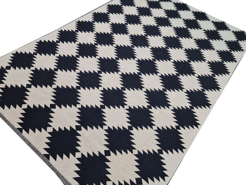 Puede incluir: Una alfombra rectangular con un patr&oacute;n repetitivo de cuadrados negros y crema. Los cuadrados tienen un borde dentado, casi en forma de estrella. La alfombra est&aacute; hecha de un material tejido y tiene una paleta de colores neutros.