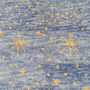 May include: Blue rug with a pattern of yellow stars. The rug has a textured appearance, with a mix of light blue and dark blue hues. The stars vary in size and are scattered across the surface, with some larger, multi-pointed stars.