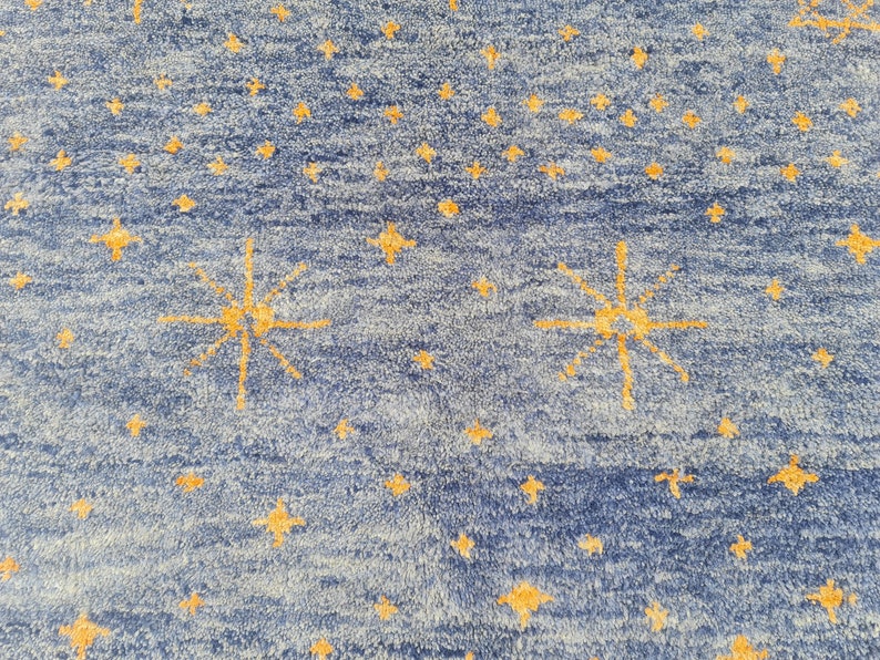 May include: Blue rug with a pattern of yellow stars. The rug has a textured appearance, with a mix of light blue and dark blue hues. The stars vary in size and are scattered across the surface, with some larger, multi-pointed stars.