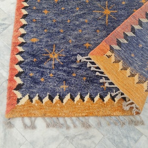 May include: A blue rug with a celestial design featuring gold starbursts and smaller stars. The rug has a border with a repeating geometric pattern in orange, black, and cream. Tassels are visible along the edge.
