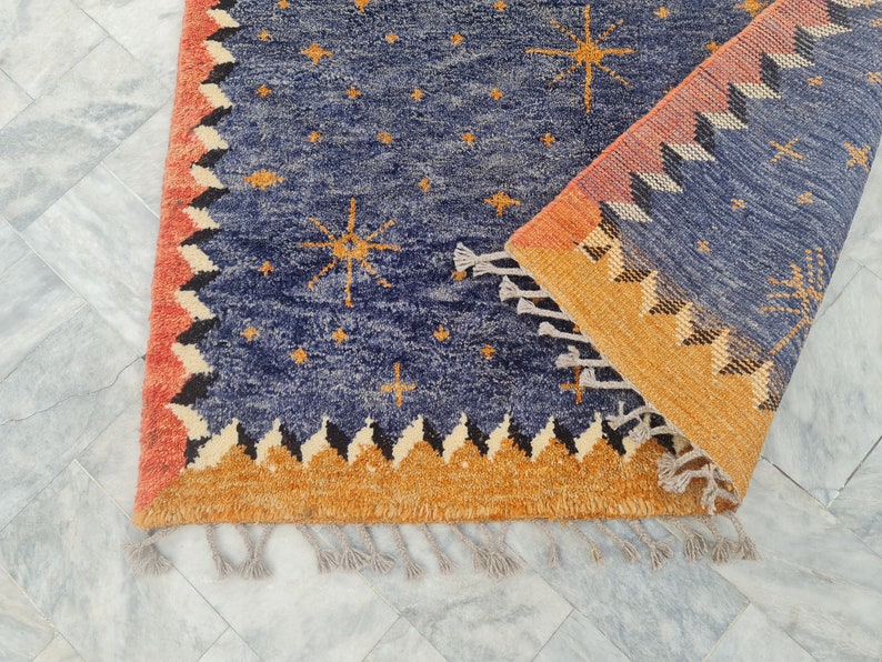 May include: A blue rug with a celestial design featuring gold starbursts and smaller stars. The rug has a border with a repeating geometric pattern in orange, black, and cream. Tassels are visible along the edge.