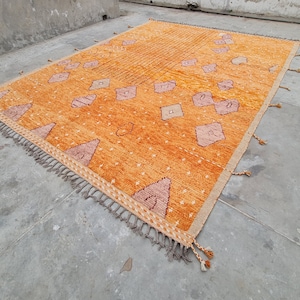Puede incluir: Una alfombra rectangular con una base de color naranja vibrante y formas geométricas dispersas en tonos rosa suave y blanco. La alfombra incluye pequeños detalles en forma de estrella blanca, una sección a rayas y flecos decorativos.