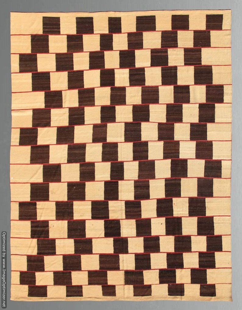 May include: A rectangular woven rug with a repeating pattern of alternating dark brown and beige blocks. Thin red lines separate the rows of blocks, creating a visually dynamic design. The rug's texture suggests a handmade quality.