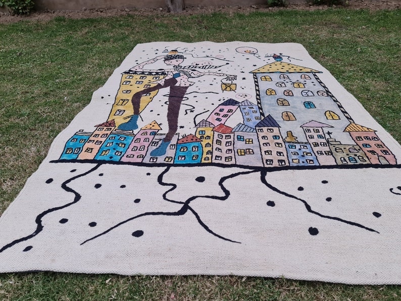 Op de afbeelding: Een groot, rechthoekig tapijt met een grillig ontwerp met een figuur en kleurrijke gebouwen. Het tapijt heeft een cr&egrave;mekleurige achtergrond met zwarte lijnen en stippen. De gebouwen zijn in verschillende kleuren geschilderd, waardoor een levendig stadsbeeld ontstaat.