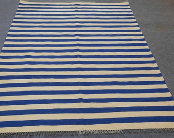 Puede incluir: Una alfombra rectangular con rayas horizontales en tonos alternos de azul y blanco. La alfombra tiene un borde con flecos y se muestra sobre una superficie gris. La alfombra es un artículo de decoración del hogar.