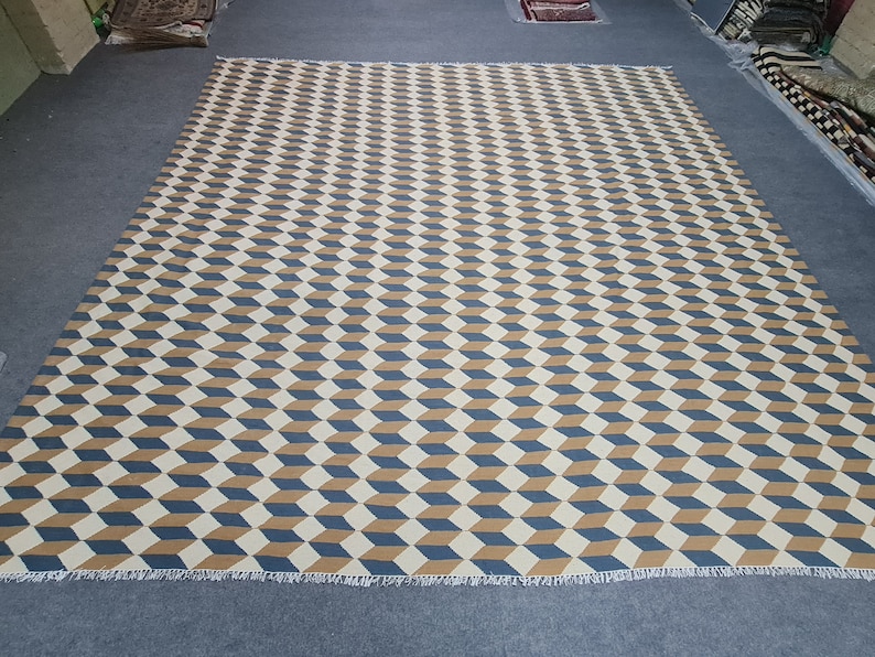 May include: Large rectangular rug with a repeating geometric pattern of three-dimensional cubes in cream, navy blue, and tan. The rug has a fringed edge and is displayed on a gray surface. The rug is suitable for a living room or bedroom.