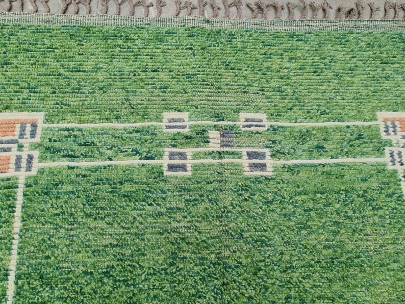 Puede incluir: Una alfombra verde y blanca con una superficie texturizada. La alfombra presenta un patr&oacute;n repetitivo de formas rectangulares en tonos de verde, blanco, beige y azul marino. El borde de la alfombra tiene flecos.
