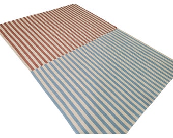 May include: A rectangular rug with a striped pattern. One half features vertical stripes in shades of red and cream, while the other half has blue and cream stripes. The rug is made of a woven material.
