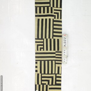 May include: A rectangular, beige and black patterned textile. The design features a patchwork of vertical and horizontal black stripes on a beige background. The textile is long and narrow, suitable for a runner or decorative piece.