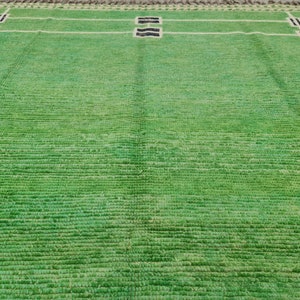 May include: A green rectangular rug with a geometric design. White lines create a grid pattern, with rectangular blocks in the corners and center. The blocks have black and brown accents. The rug has a textured surface.