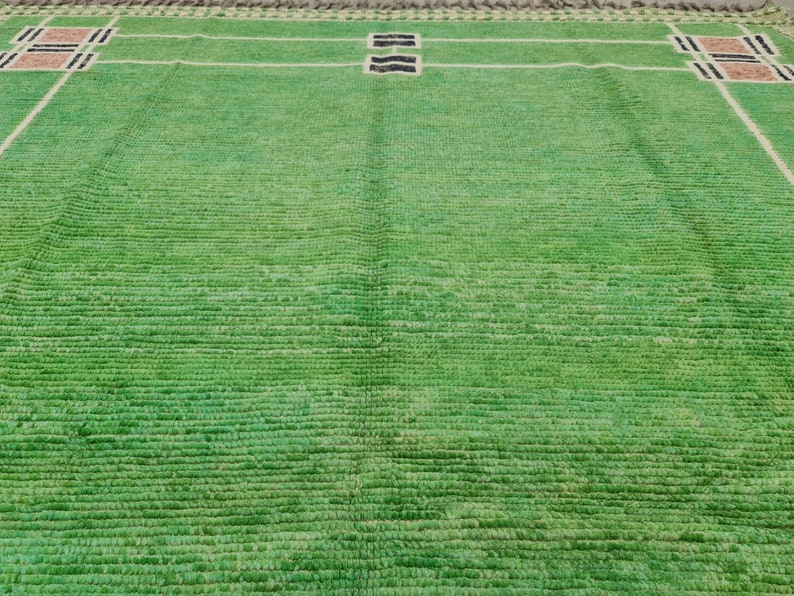 May include: A green rectangular rug with a geometric design. White lines create a grid pattern, with rectangular blocks in the corners and center. The blocks have black and brown accents. The rug has a textured surface.