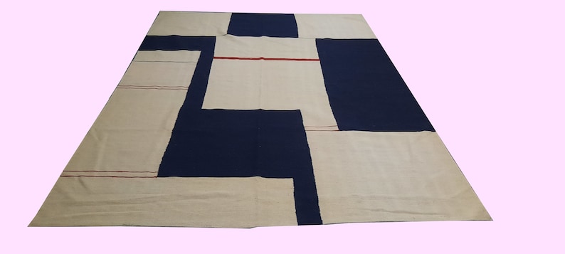 Op de afbeelding: Een groot, rechthoekig tapijt met een geometrisch ontwerp. Het tapijt heeft een patchworkpatroon van marineblauwe, gebroken witte en beige secties. Dunne rode lijnen accentueren enkele van de gebroken witte panelen. Geschikt voor een woon- of slaapkamer.