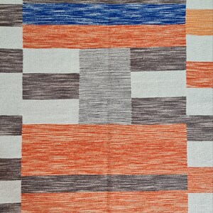 May include: A woven rug with a geometric pattern. The design features alternating blocks of color, including orange, blue, gray, and white. The rug's texture appears to be made of woven fibers, creating a textured surface. The overall aesthetic is modern and abstract.