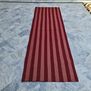May include: A rectangular rug with vertical stripes in shades of red and pink. The rug is placed on a light-colored tiled floor. The rug's design features alternating stripes of varying widths, creating a visually appealing pattern.