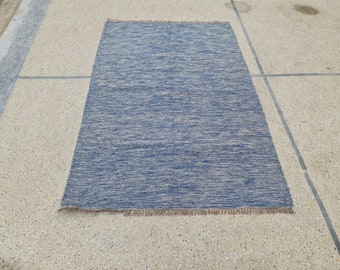 Puede incluir: Una alfombra rectangular tejida en azul y beige con un borde con flecos. La alfombra tiene un aspecto texturizado con una mezcla de hilos azules y beige. La alfombra mide aproximadamente 122 cm de largo y 76 cm de ancho.