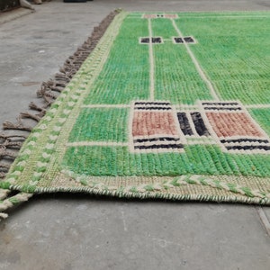 May include: A green and beige rectangular rug with a geometric design. The rug features a light green field with white lines and rectangular accents in shades of brown and black. The edges have a fringe.