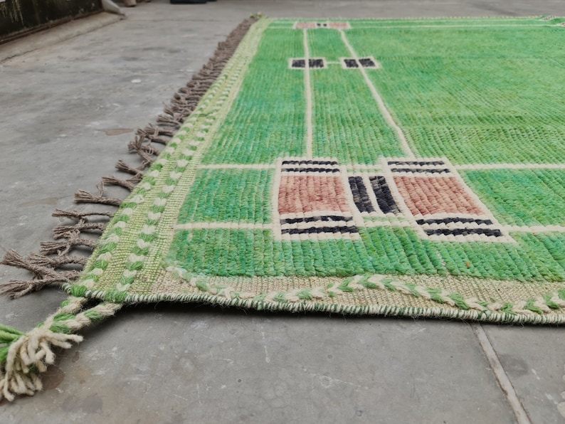 May include: A green and beige rectangular rug with a geometric design. The rug features a light green field with white lines and rectangular accents in shades of brown and black. The edges have a fringe.
