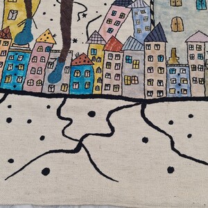 Op de afbeelding: Een grillig, handgetekend tapijt met een kleurrijke stadsgezicht met gebouwen in verschillende kleuren, waaronder blauw, roze en geel. Het ontwerp bevat zwarte contouren en een cr&egrave;mekleurige achtergrond.