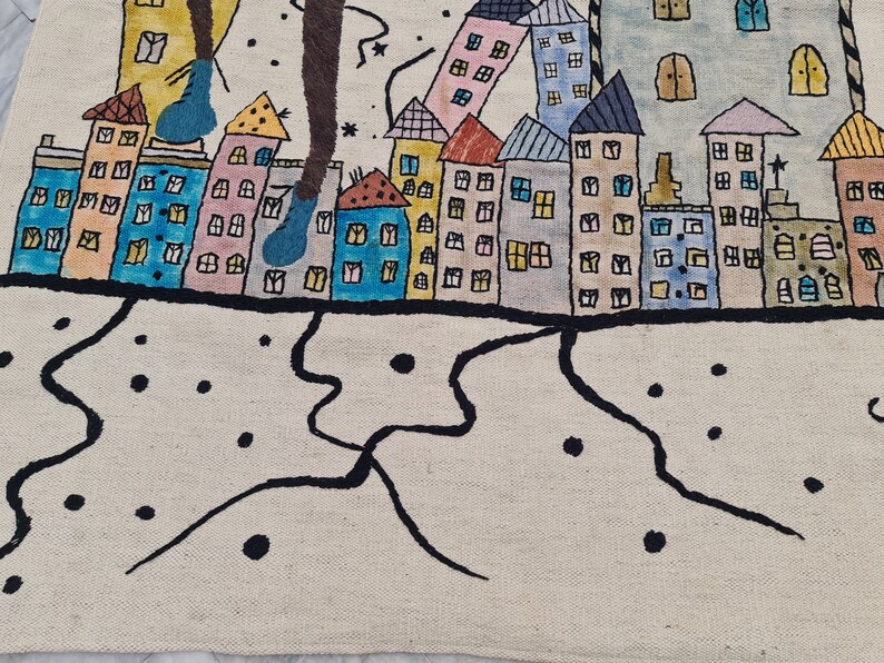 Op de afbeelding: Een grillig, handgetekend tapijt met een kleurrijke stadsgezicht met gebouwen in verschillende kleuren, waaronder blauw, roze en geel. Het ontwerp bevat zwarte contouren en een cr&egrave;mekleurige achtergrond.