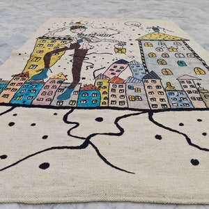 Op de afbeelding: Een cr&egrave;mekleurig tapijt met een grillige stadsgezicht. Het ontwerp toont kleurrijke gebouwen, een figuur met uitgestrekte armen en zwarte abstracte lijnen. Het tapijt is gemaakt van een geweven materiaal.