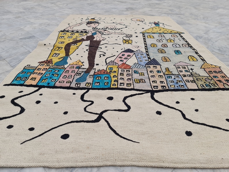 Op de afbeelding: Een cr&egrave;mekleurig tapijt met een grillige stadsgezicht. Het ontwerp toont kleurrijke gebouwen, een figuur met uitgestrekte armen en zwarte abstracte lijnen. Het tapijt is gemaakt van een geweven materiaal.