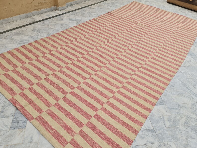May include: A large rectangular rug with a repeating pattern of alternating red and beige stripes. The stripes run horizontally and vertically, creating a checkered design. The rug is made of a woven material.