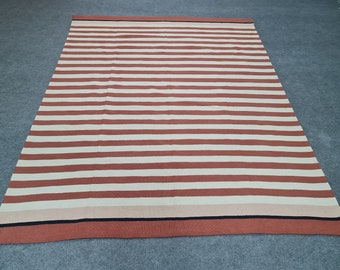 Könnte beinhalten: Ein rechteckiger Teppich mit horizontalen Streifen in Rot- und Weißtönen. Der Teppich hat einen Rand mit einem schwarzen Streifen. Der Teppich besteht aus einem gewebten Material und ist für den Innenbereich geeignet.