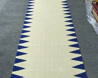 May include: A long, rectangular rug with a cream-colored center and a border of blue triangles. The rug is made of woven material and is displayed on a gray floor. The rug's design is symmetrical, with the blue triangles pointing inwards.