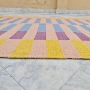 May include: A rectangular woven rug with a colorful geometric pattern. The rug features alternating blocks of yellow, pink, blue, and purple. The rug is made of a thick, textured material and is suitable for indoor use.