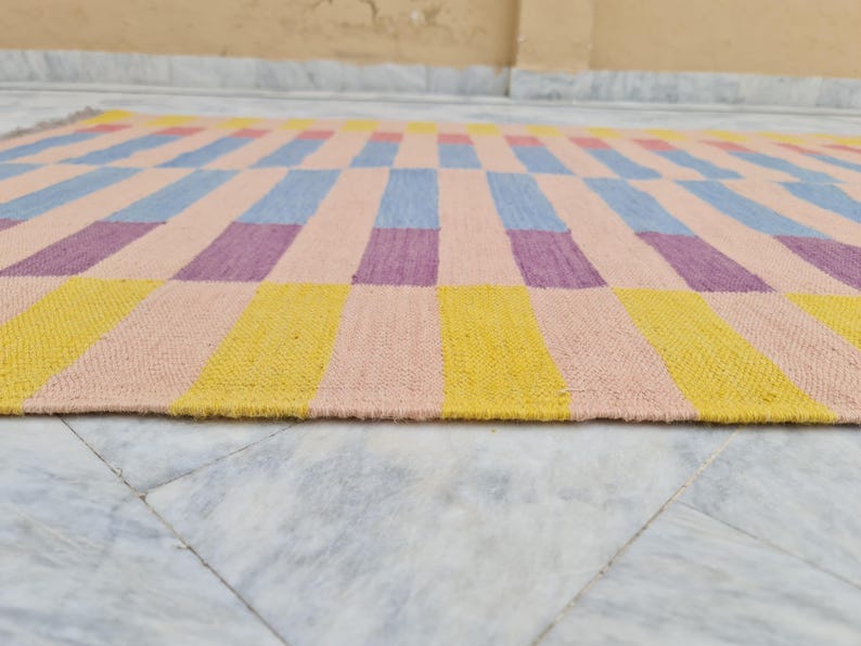 May include: A rectangular woven rug with a colorful geometric pattern. The rug features alternating blocks of yellow, pink, blue, and purple. The rug is made of a thick, textured material and is suitable for indoor use.