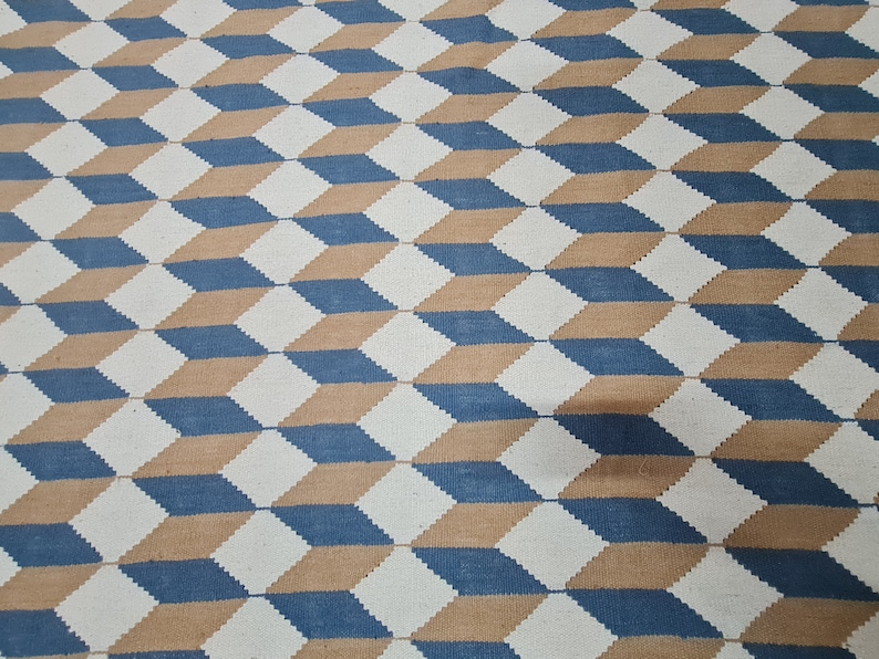 May include: Geometric patterned rug featuring a repeating 3D cube design. The rug's pattern uses a combination of white, blue, and tan colors. The design creates an optical illusion of depth and dimension, suitable for home decor.