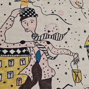 Op de afbeelding: Geborduurd kunstwerk dat een fantasierijke sc&egrave;ne toont met een figuur in een gestippeld shirt en geruite hoed, rennend langs kleurrijke gebouwen. Het kunstwerk heeft een lichte achtergrond met sterren en zwevende objecten, wat een speelse, dromerige sfeer cre&euml;ert.