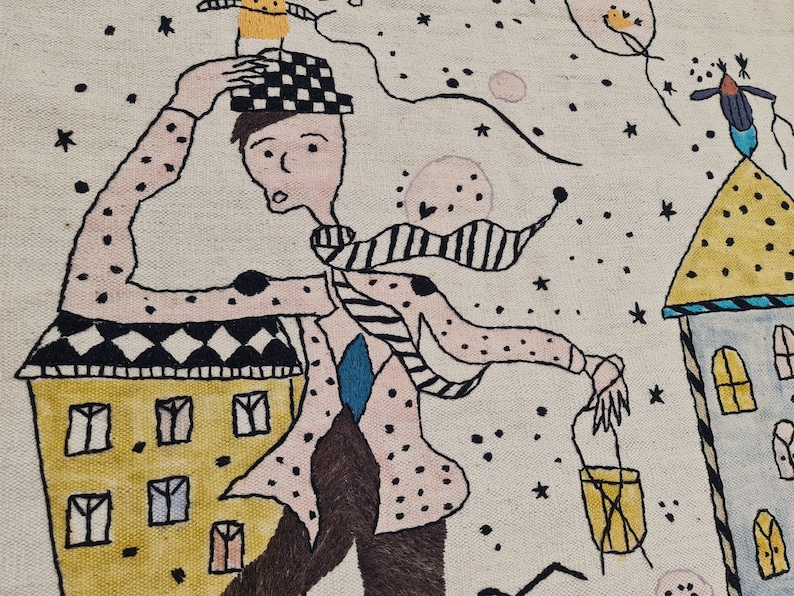 Op de afbeelding: Geborduurd kunstwerk dat een fantasierijke sc&egrave;ne toont met een figuur in een gestippeld shirt en geruite hoed, rennend langs kleurrijke gebouwen. Het kunstwerk heeft een lichte achtergrond met sterren en zwevende objecten, wat een speelse, dromerige sfeer cre&euml;ert.