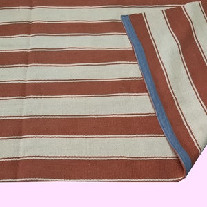May include: A striped rug with alternating bands of rust-colored and off-white. The rug has a blue border on one side, and is partially folded over, revealing the reverse side with the same pattern. The rug is likely made of woven fabric.