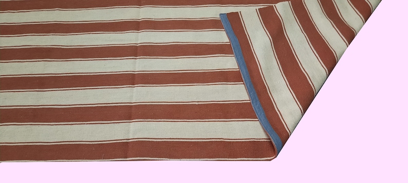 May include: A striped rug with alternating bands of rust-colored and off-white. The rug has a blue border on one side, and is partially folded over, revealing the reverse side with the same pattern. The rug is likely made of woven fabric.